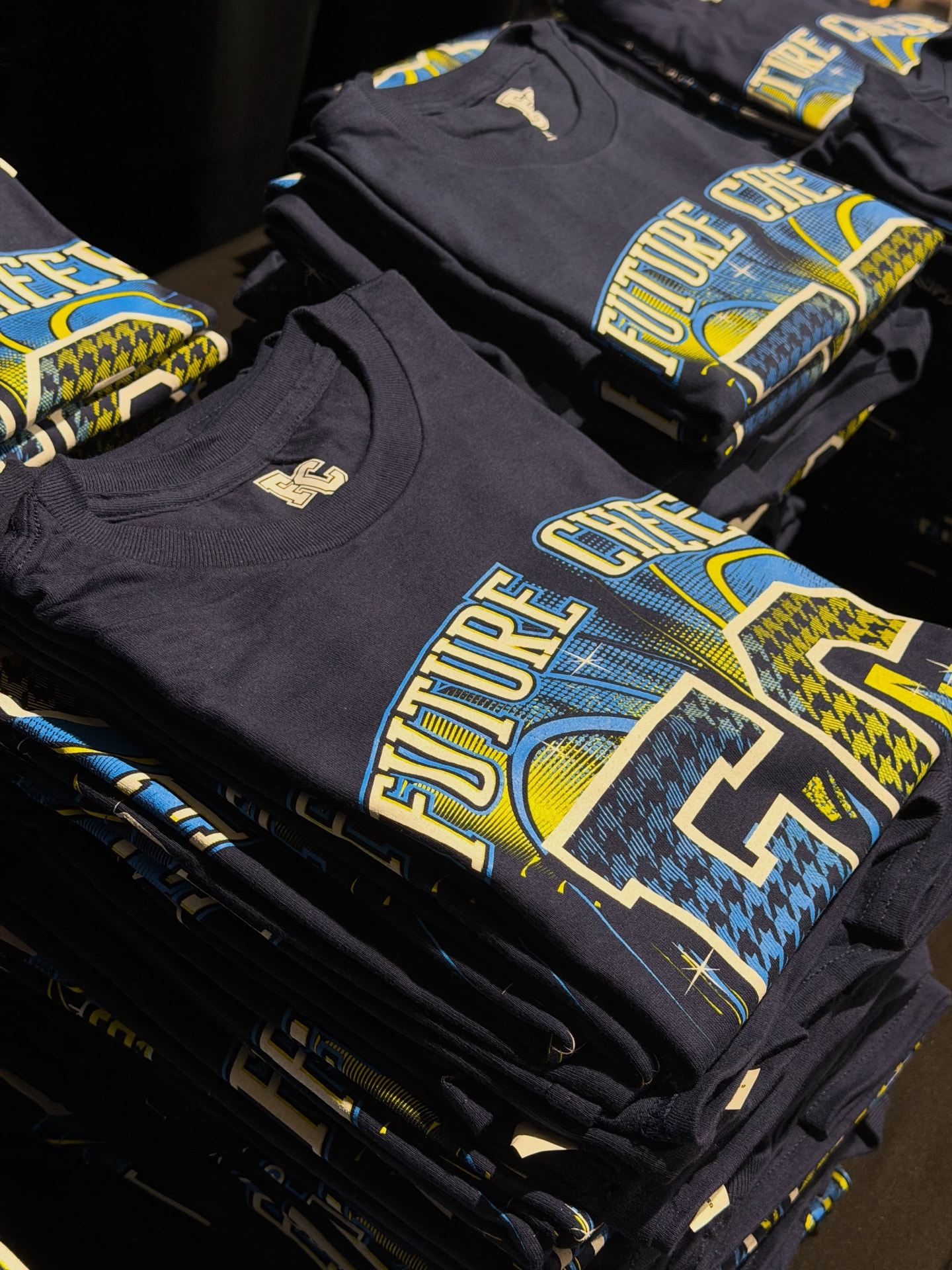 Stacks of folded navy Future Cheer t-shirts printed with blue and gold graphics